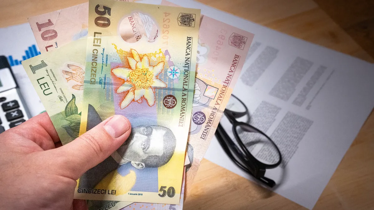 bancnote românești (lei) ținute în mână deasupra unei mese pe care se află un calculator, documente și o pereche de ochelari
