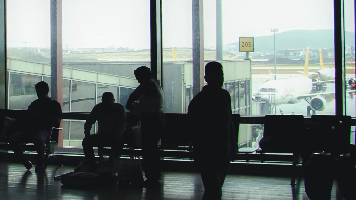 Siluete ale unor călători care așteaptă în terminalul aeroportului din Ankara. Surprinde atmosfera de așteptare și de călătorie.
