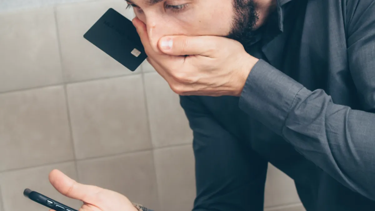 Un bărbat se uită îngrozit la smartphone în timp ce ține un card bancar în mână